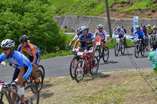 コンクリートの坂道を、多くの選手たちがマウンテンバイクで一列になって駆け上っているレース中の写真