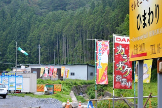 緑の山あいにのぼり旗が何本も立つプレハブの建物が建っている写真