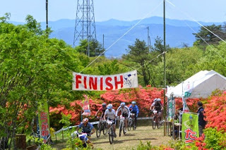 「FINISH」と書かれた横断幕の下、赤いツツジと山々を背景に複数の選手が次々とゴールラインを越えてくる瞬間の、たつがねMTB大会のフィニッシュ地点の写真