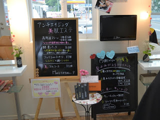 店舗の壁際に設置された黒板に「アンチエイジング 美肌エステ」と書かれ、サービス内容と料金が案内されている写真