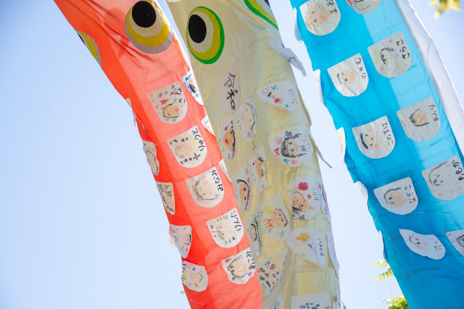 赤・黄色・青の鯉のぼりが風に揺れ、それぞれの鱗に子どもたちの顔の絵や名前が貼られている様子を近くから写した写真