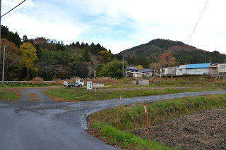秋色に染まった木々が広がる山に囲まれた田畑の看板の側にトラックが停まり、住宅が広がっている写真