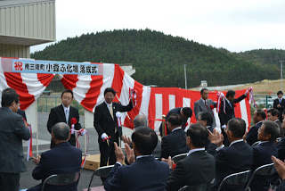 来賓の方が壇上で赤と白の幕の前に立ち、テープカットをしているところを、スーツ姿の多くの出席者が椅子に座って拍手して見守っている屋外式典の写真