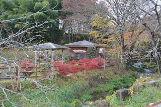 紅葉が始まりかけた木々に囲まれた小さな東屋が二つ並んでおり、前には赤い低木が鮮やかに色づき、小川が静かに流れている秋の山間の風景の写真