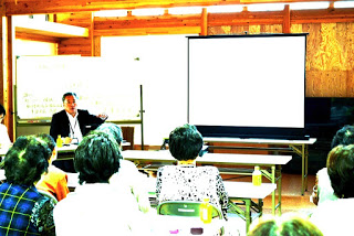 木造の明るい室内で、スーツ姿の講師がスクリーンを使って講義を行い、高齢の参加者たちが熱心に耳を傾けている様子の写真