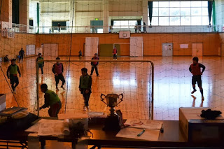 体育館の室内で複数の男性がフットサルをしており、ネット越しにゴールとボールが見える様子の写真