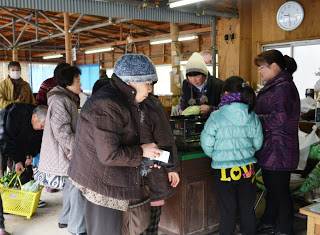 木の梁が見える屋内の店舗で、冬服を着た人々や黄色い買い物かごを持つ男性などがレジを待ちをしている買い物の様子が写されている写真