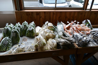 木の板張りの壁と窓が見える店内で、陳列棚にインゲン、ジャガイモ、ニンジン、ナスなどの新鮮な野菜がそれぞれ透明な袋に入れられて並べられている写真