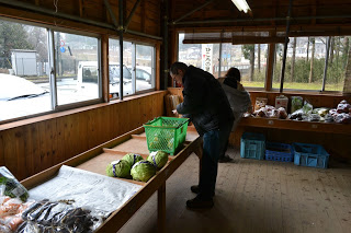 木の壁と大きな窓のある小さな店舗内で、テーブルの上に置かれたキャベツなどの野菜が入った緑のかごのそばに一人が立ち、奥の陳列棚にもう一人お客さんがいる、野菜や日用品を販売する店内の写真