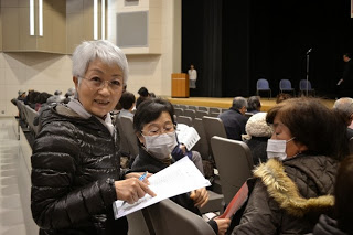 多くの人がコートやマスクを着けて椅子に座っている講堂で、白髪の女性が前を向きながら紙を持って話しかけている、講演会の開演前の様子の写真