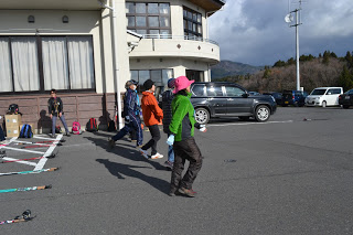 背景には車や木々が見える建物前の駐車場の地面にはスキーポールやリュックサックが置かれていて、建物の近くに複数の参加者が横一列に並び、ウォーミングアップを行っている屋外活動の準備をしている様子が写っている写真