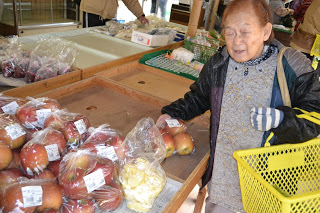 リンゴなどの果物がそれぞれビニール袋に入れて並べられており、女性客が黄色い買い物かごを持って果物を選んでいる様子の写真