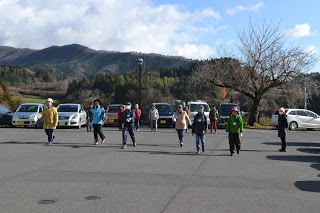 背景には山や木々、そして部分的に雲がかかった空が広がっている屋外の駐車場で、複数の参加者が歩いてウォーミングアップを行う様子が写っている写真