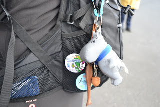 黒のリュックサックのアップ写真で、イラストと文字が描かれた缶バッジと青いデザインのバッジが2つ付けられており、青いマフラーをつけたムーミンのぬいぐるみ型キーホルダーがぶら下がっている写真