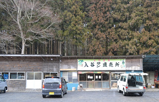 「入谷サン直売所」と書かれた看板が掲げられた小さな木造の建物の前に、黒と白の車が停まっている直売所の写真
