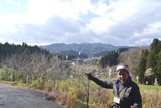 山や木々が広がる風景の中で、ジャケットを着て首に名札をかけ、ウォーキングポールを持って手を伸ばし、右側を指さしている女性が屋外に立っている様子の写真