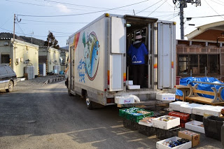 トラックの荷台で商品を整理している人がおり、その周囲には整然と並べられた複数の箱や商品が置かれている様子が写っている写真