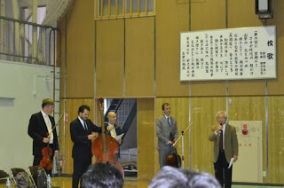 体育館のような広い室内で、4人の男性がそれぞれ楽器を持って立っており、マイクを持ち右側に立つ男性が話をしている様子の写真