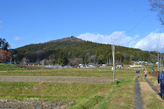 青空と雲が広がる中、山のふもとには木々、田畑や家々が広がり、右側の小道を数人の参加者が歩いている写真