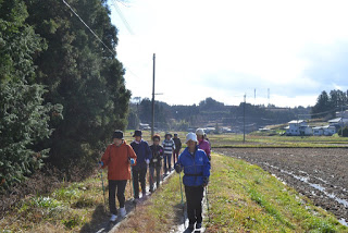 道の片側には木々、もう片側には畑が広がり、背景にはさらに畑や家々がある細い道を、屋外用の服装を着て一部がウォーキングポールを使いながら歩いている複数の参加者が写っている写真