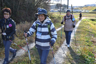 草地や木々、遠くに建物が見える屋外で、複数の参加者がウォーキングポールを使いながらあぜ道を歩いている様子が写っている写真