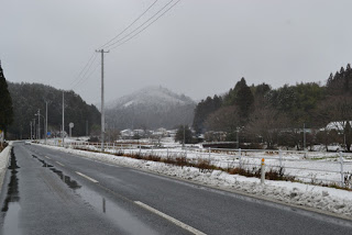 山頂に雪が積もった山が霞んで見える方向に向かって片側一車線の道路がまっすぐに伸び、その両側には雪に覆われた木々や家々が静かに佇む冬の風景が広がっている写真