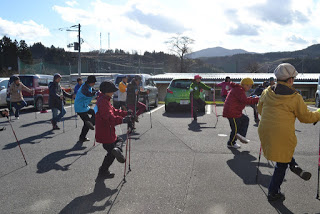 木々と山々が背景に広がる駐車場で、冬服を着た複数の参加者がウォーキングポールを使いながら片足をあげる体操を行っている様子が写っている写真