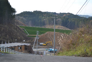 坂道の先にある造成地でショベルカーなどの重機が作業しており、その周囲を緑の法面と杉林が囲んでいる造成地の工事を離れたところから写した写真