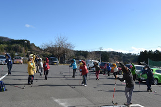 青空の下、木々や丘が背景に広がる開けた駐車場で、冬服を着た複数の参加者がウォーキングポールを使いながら体操をしている様子が写っている写真