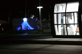夜の暗がりの道路沿いに、右側には大きな窓から明るい光が漏れる建物があり、左奥には青いライトで飾られたクリスマスツリーが見える写真