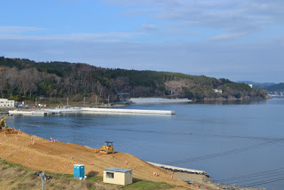 重機が作業している海岸沿いの造成地の先に穏やかな海が広がり、その奥に防波堤と入江が見える海辺の工事の写真