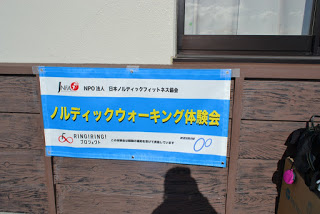 「NPO法人 日本ノルディックウォークフィットネス協会 ノルディックウォーキング体験会」と書かれた青と白の旗が、建物の木の壁に取り付けられている写真