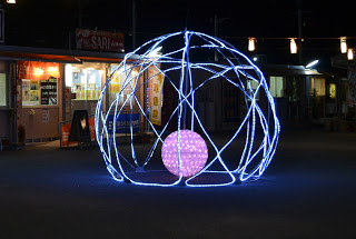 夜の屋外に設置された光のアート作品で、青いLEDライトで構成されたワイヤーフレーム状の大きな球体の中に、ピンク色に光る小さな球体が浮かんでいるイルミネーションの写真