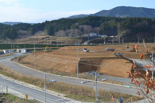 背景に山々が広がるカーブの道路の内側に造成された広い土の台地があり、重機や車両が点在し工事中の写真