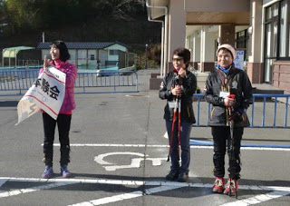 背景には青い金属製の柵が写り建物の前の駐車場に3人の人物が立っており、左の人物は「NW体験会」と書かれた旗を持ち、他の2人はウォーキング用の杖を持って屋外活動の服装をして並んでいる写真