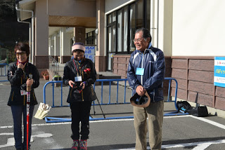 青いバリケードが立てられた建物入り口前の駐車場で、男性1人と女性2人が、ウォーキングポールを持って笑顔で立っている写真
