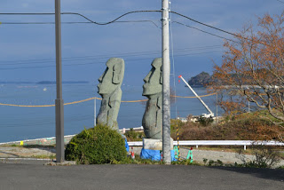 海を背景に並ぶ2体の大きなモアイ像が立っており、奥にはクレーンが見える港の様子が広がっている写真