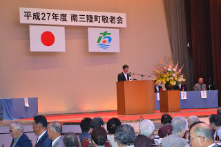 「平成27年度 南三陸町敬老会」の式典で、国旗と南三陸町町旗の下、演台に立った町長が挨拶をしている写真