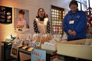 壁にポスターが貼られている屋内の場所で、2人の人物がテーブルの後ろに立っており、テーブルの上には値札が付いた複数の包装された食品が並べられている写真