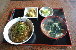 黒いお盆の上に、ご飯の上にめかぶが乗った丼、海鮮ハット汁、牡蠣のてんぷらが2つ乗った小皿、漬物が盛られた小皿が並んだ写真