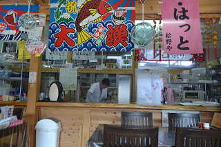 天井の梁に飾られた「大漁」と書かれた大きな大漁旗やピンクの旗、ガラスの浮き玉などの後ろに調理服を着た男性が調理をしている様子が見える写真