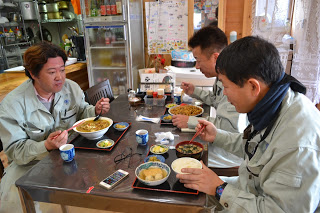 テーブル席に座った作業着姿の3人の男性が、それぞれ定食や麺類などを食べている写真