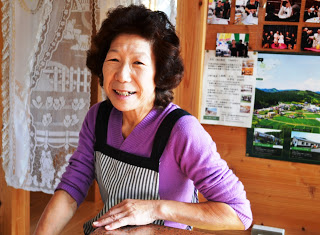 レースの暖簾が掛けられ、壁に複数の写真などが貼られた店内で、テーブルに手を置いたエプロン姿の女性が笑顔で写っている写真