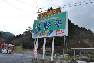 山や木々に囲まれた中の大きな道路沿いに「峠の茶や・松野や」営業時間「11時~2時」と書かれた看板が立っており、道路の反対側には赤い自動販売機が写っている写真