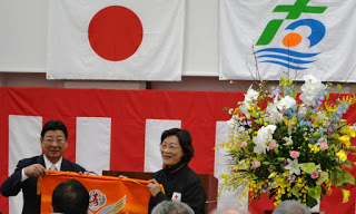 紅白幕や国旗と町旗が掲げられた前で、豪華な花が飾られた横で佐藤町長と女性がオレンジ色の大漁旗を広げている場面を写した式典の写真