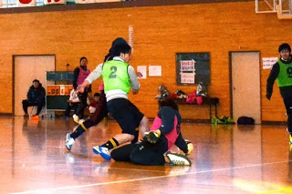 ピンクのビブスを着たゴールキーパーがひざまずいてボールをキャッチしようとしているところに、黄緑のビブスを着た選手が近づいている瞬間の写真