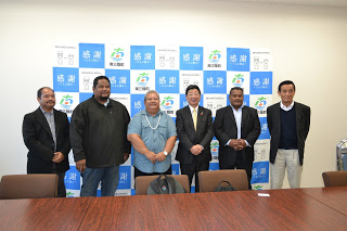 南三陸町のバックパネルの前で、佐藤町長を中心にパラオ共和国の関係者等6人の男性が並んで立っている記念写真