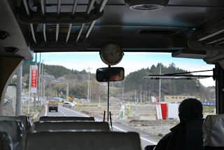 バスの車内後方からフロント方向を写しており、前方のフロントガラス越しに道路と建物、山が見え、運転手と乗客の後ろ姿が写っているバス移動中の車内の写真