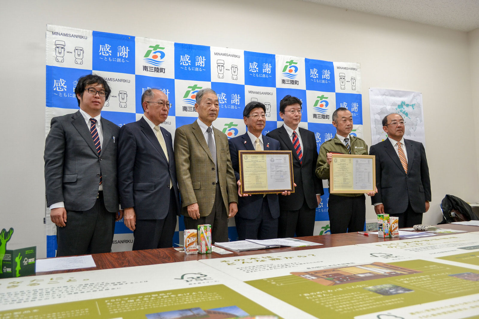 南三陸町のバックパネルの前で、スーツ姿の男性たち7人が並び、2人が額入りの証明書を掲げて記念撮影している写真