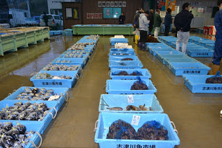 屋内の魚市場で、複数の青い容器にさまざまな種類の海産物が入って並べられており、容器にはラベルが付けられ、数人の人々が市場内を歩いたり立ち止まったりしている様子が写された、魚市場の写真